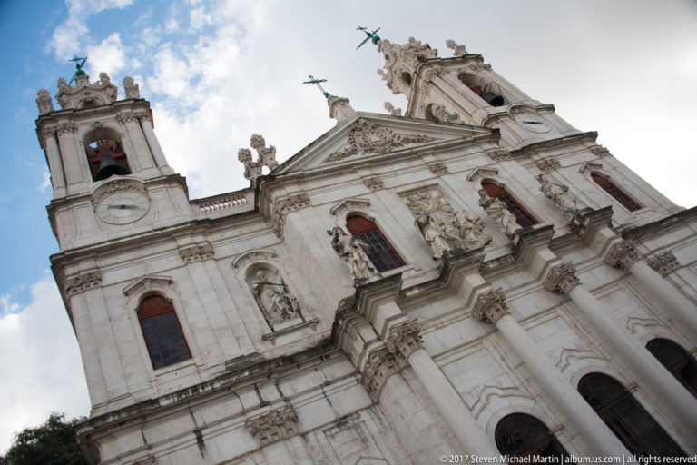 Low-angle view of the Basílica da Estrela façade with twin bell towers, saint statues, clocks, columns, and central pediment.