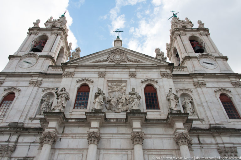 Low-angle view of the Basílica da Estrela façade with twin bell towers, clocks, saint statues, and carved pale-stone ornament.