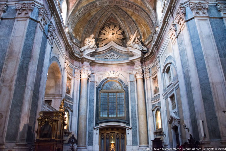 Side interior of the Basílica da Estrela with tall window, organ case, marble wall surfaces, sculpted angels, and radiant emblem.