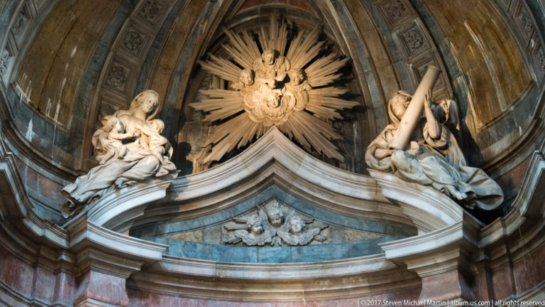 Two white sculpted angels flanking a radiant sacred emblem above a curved marble pediment in the Basílica da Estrela.