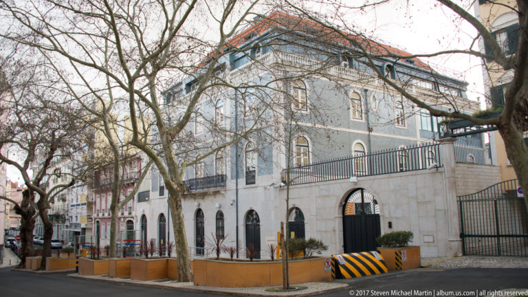 Blue-gray historic corner building with arched ground-floor openings, wrought-iron railings, red-tiled roof, and leafless street trees.