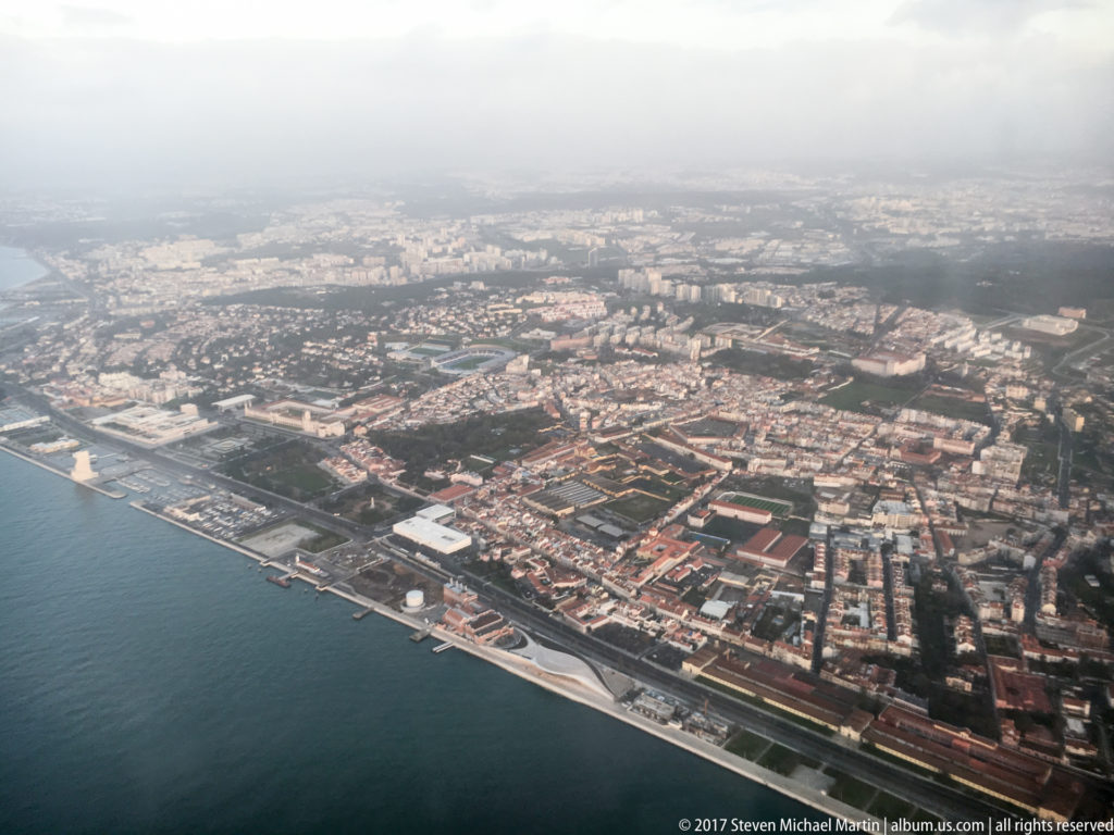 Aerial view over dense Lisbon neighborhoods and a long developed waterfront beside broad blue-gray water.