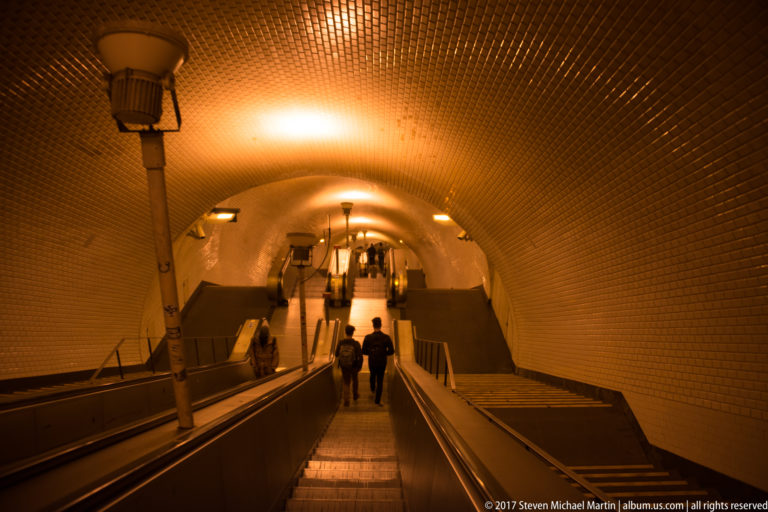 Long escalators descending through a warmly lit vaulted and tiled Lisbon Metro passage with several passengers.