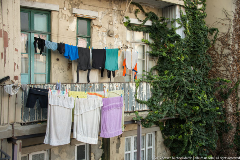 Laundry in several colors hangs from lines and balcony rails on a weathered residential building partly covered by vines.