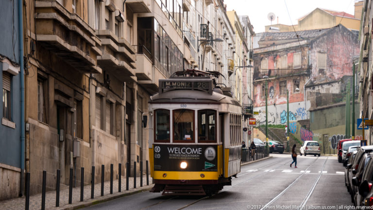 Traditional yellow-and-white Lisbon tram approaching on embedded street rails between tall buildings.