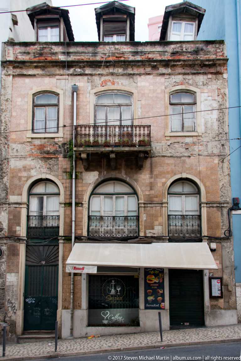 Weathered multi-story Lisbon façade with arched windows, iron balconies, and storefronts at street level.