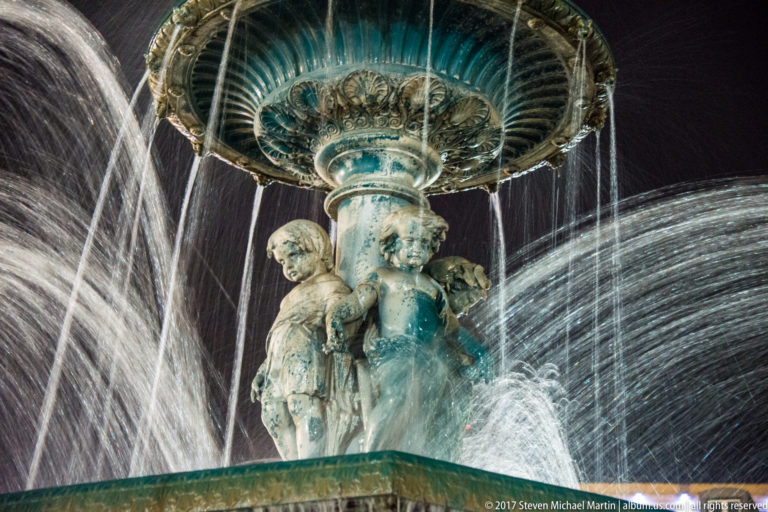 Close view of sculpted figures beneath a fountain basin, surrounded by streams and sprays of water at night.