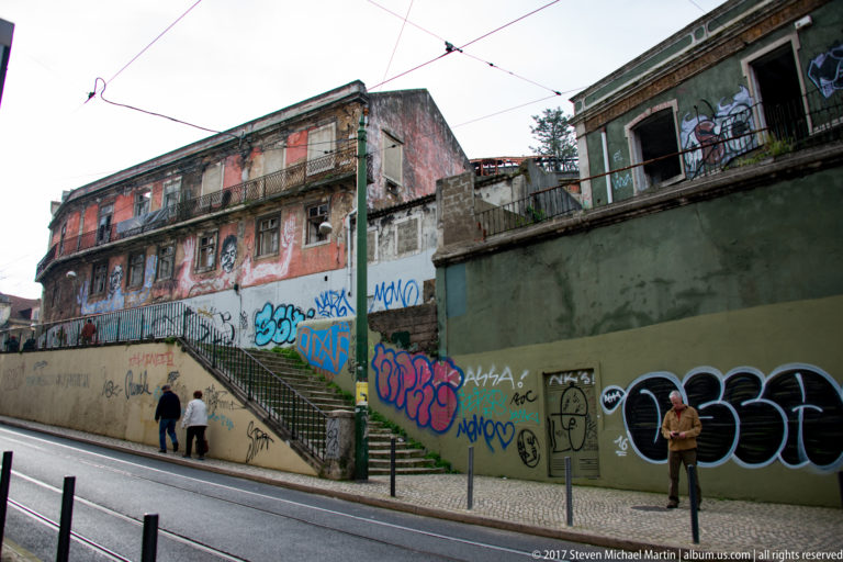 Sloping Lisbon street with graffiti-covered retaining walls, stairs, deteriorated buildings, pedestrians, and overhead tram wires.