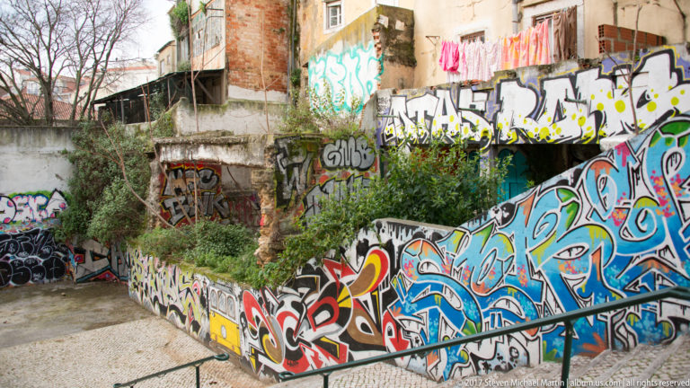 Overgrown vacant lot and ruined walls covered in colorful graffiti, with laundry hanging from a building above.