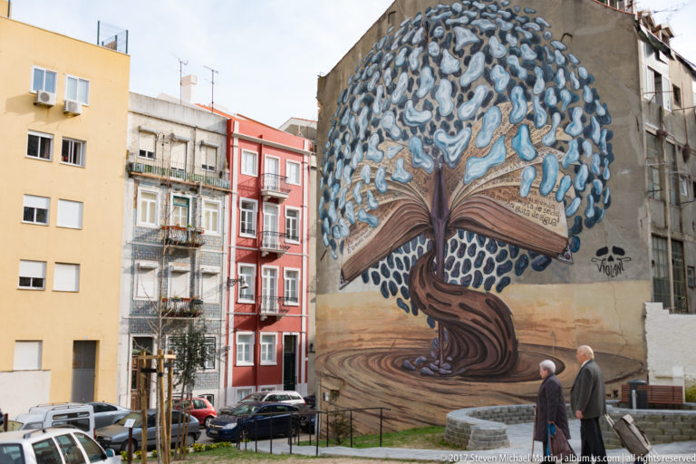 Large mural of a twisting tree with blue-gray foliage covering a tall apartment wall, with two pedestrians below.