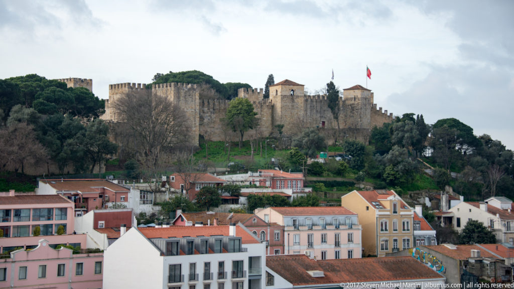 Crenellated stone walls and towers of São Jorge Castle on a wooded hill above red-roofed Lisbon buildings.