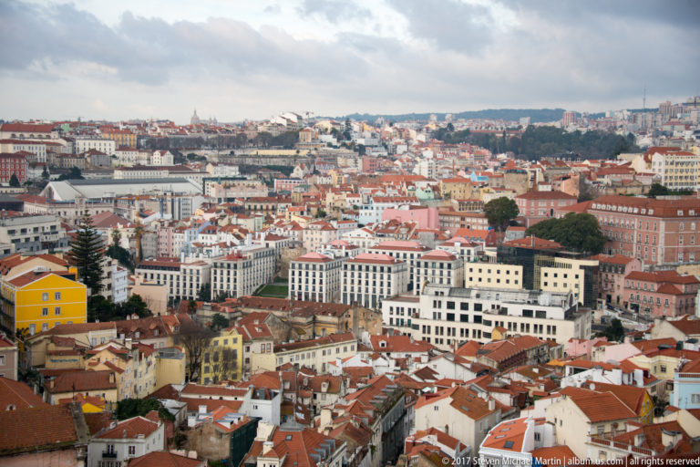 Elevated panorama over dense central Lisbon with red-tiled roofs, pale buildings, and distant neighborhoods under cloudy sky.