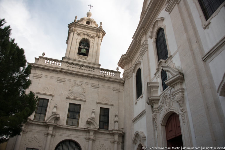 Close upward view of adjoining pale stone church façades with an ornate portal, arched windows, balustrade, and bell tower.