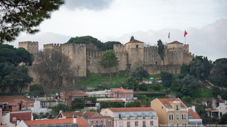 São Jorge Castle with crenellated walls and towers on a green hill above pastel buildings and red roofs.