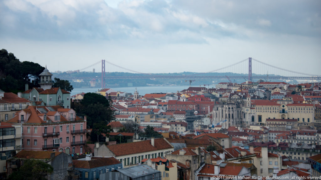 Lisbon rooftops in the foreground with the Tagus River and red 25 de Abril suspension bridge in the distance.