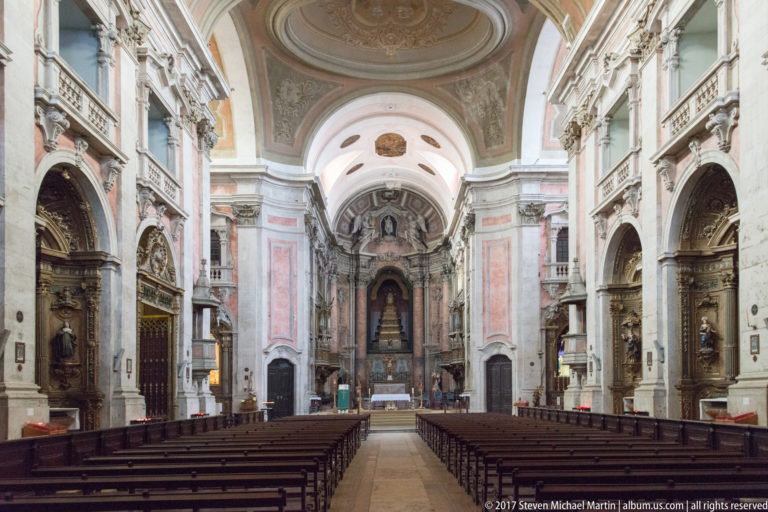 Symmetrical church nave with wooden pews, gilded side chapels, colored marble, painted vault, and distant high altar.