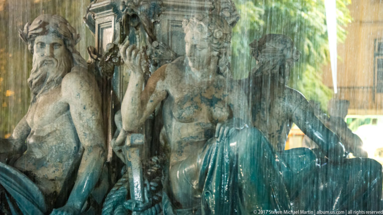 Weathered stone fountain figures seen close up behind cascading water and wet sculptural surfaces.