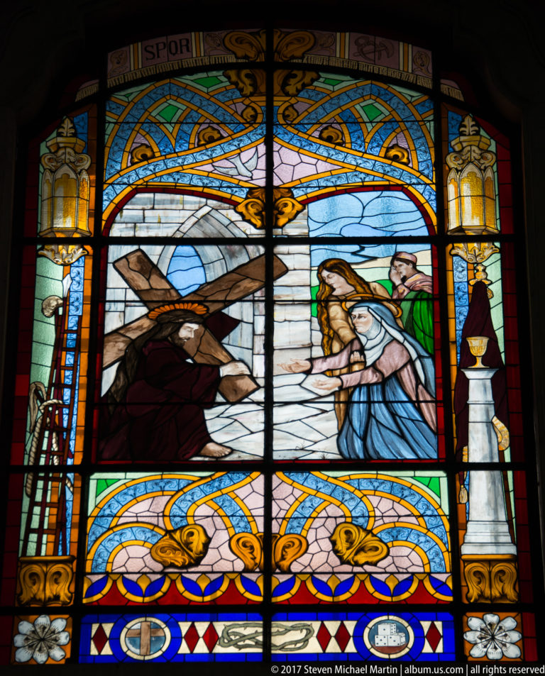 Religious stained-glass window with several robed figures, blue sky, decorative borders, and heraldic motifs below.