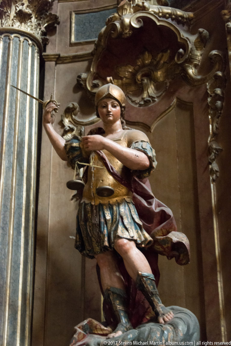 Polychrome female saint statue holding balance scales inside a carved and gilded church niche.