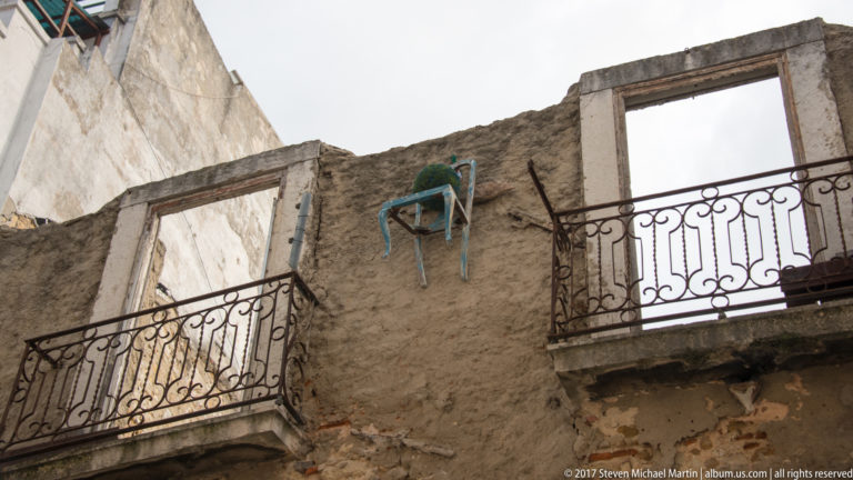 Roofless weathered masonry wall with two iron balconies, empty window openings, and a chair lodged high above.
