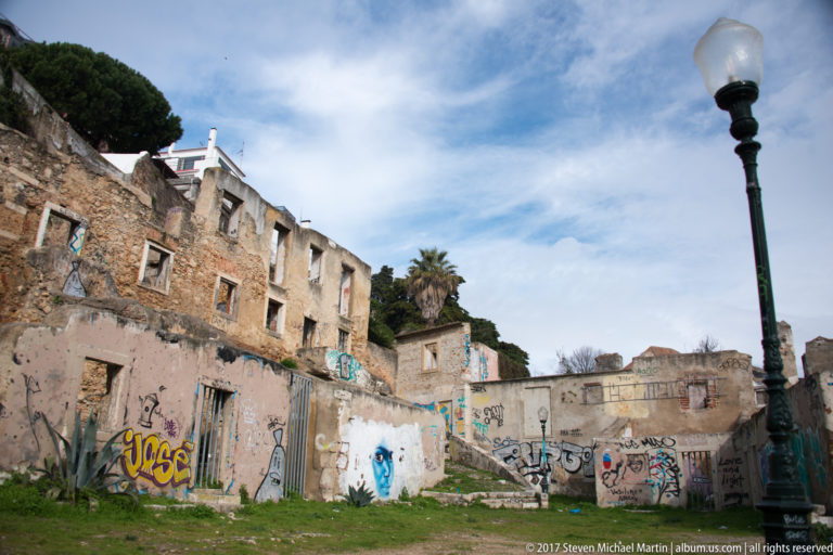 Graffiti-covered ruined buildings beside open ground, vegetation, and a tall traditional streetlamp under a blue sky.
