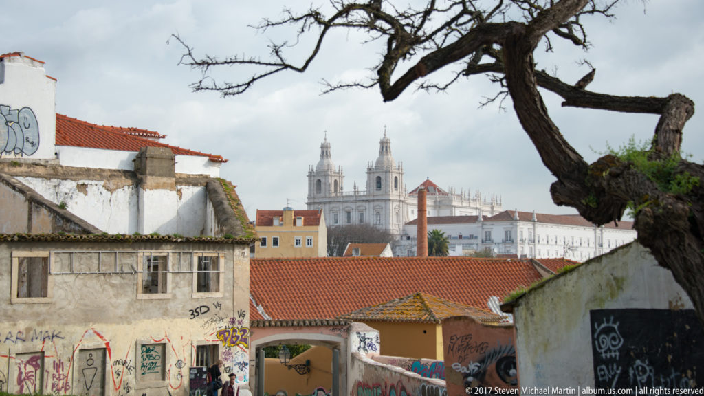 São Vicente de Fora's twin towers rising beyond red-tiled Lisbon roofs, framed by a large tree and weathered walls.
