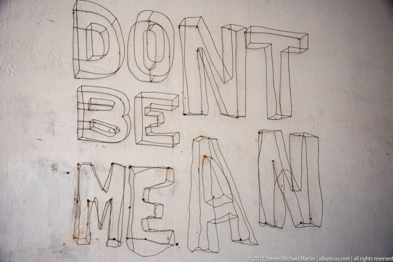 Pale wall marked with large outlined graffiti letters reading 'DONT BE MEAN.'