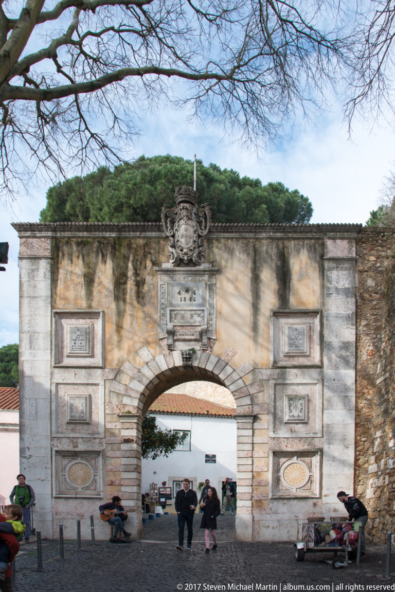 Stone arched gateway at São Jorge Castle with carved panels, royal arms, inscription tablet, and visitors passing beneath.