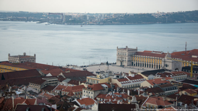 Elevated view over red Lisbon roofs to Praça do Comércio and the broad Tagus River beyond.