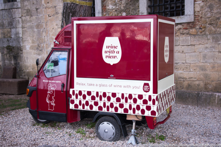 Red three-wheeled mobile wine stand labeled 'wine with a view' parked beside an old stone wall.