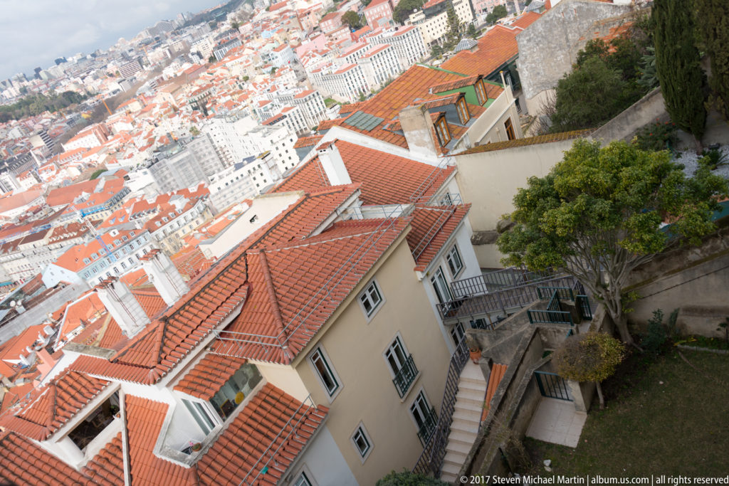 Steep hillside of pale houses and terracotta roofs with stairs, walls, and trees descending through historic Lisbon.