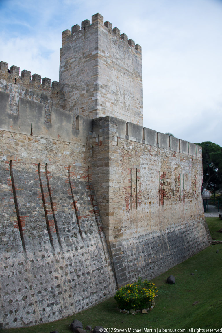 Stone battlement and square crenellated tower of São Jorge Castle above a sloping lawn.
