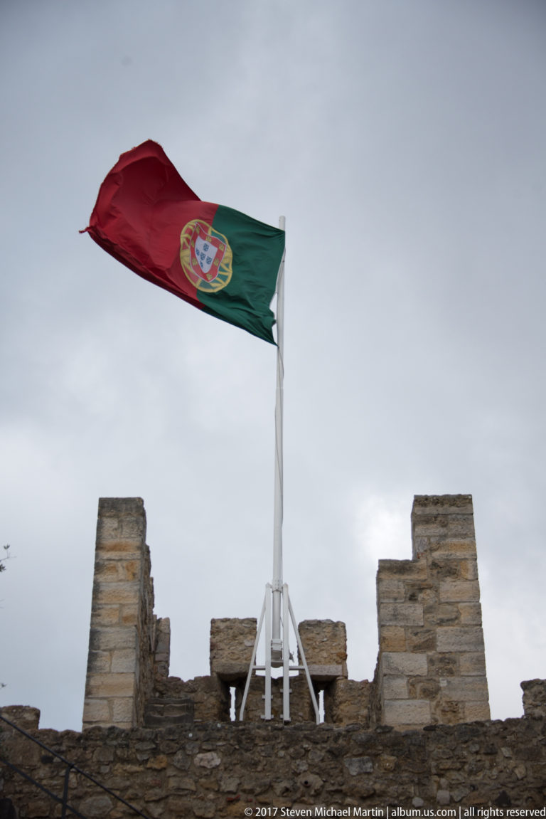 Portuguese flag flying above rough stone battlements of São Jorge Castle beneath a gray sky.