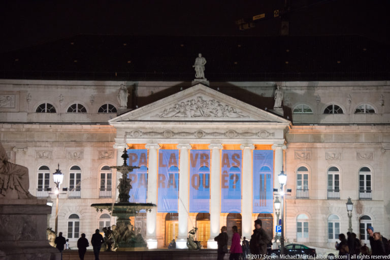 Neoclassical Teatro Nacional D. Maria II façade at night with a large text projection across its columned entrance.