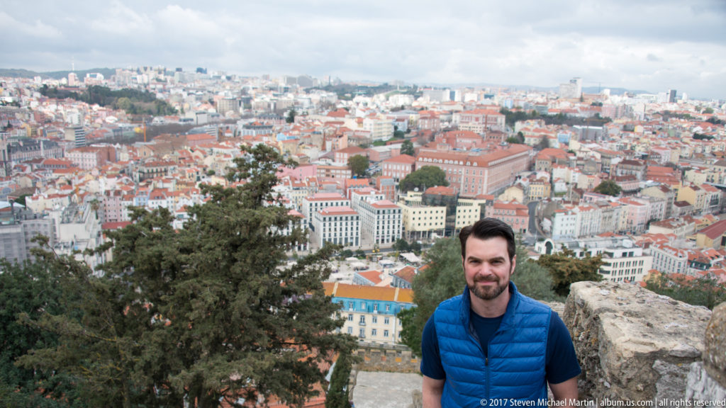 Man in a blue vest at a stone castle viewpoint with central Lisbon filling the background.