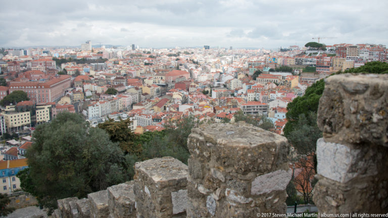 Stone castle crenellations in the foreground overlooking a broad panorama of Lisbon's red-roofed neighborhoods.