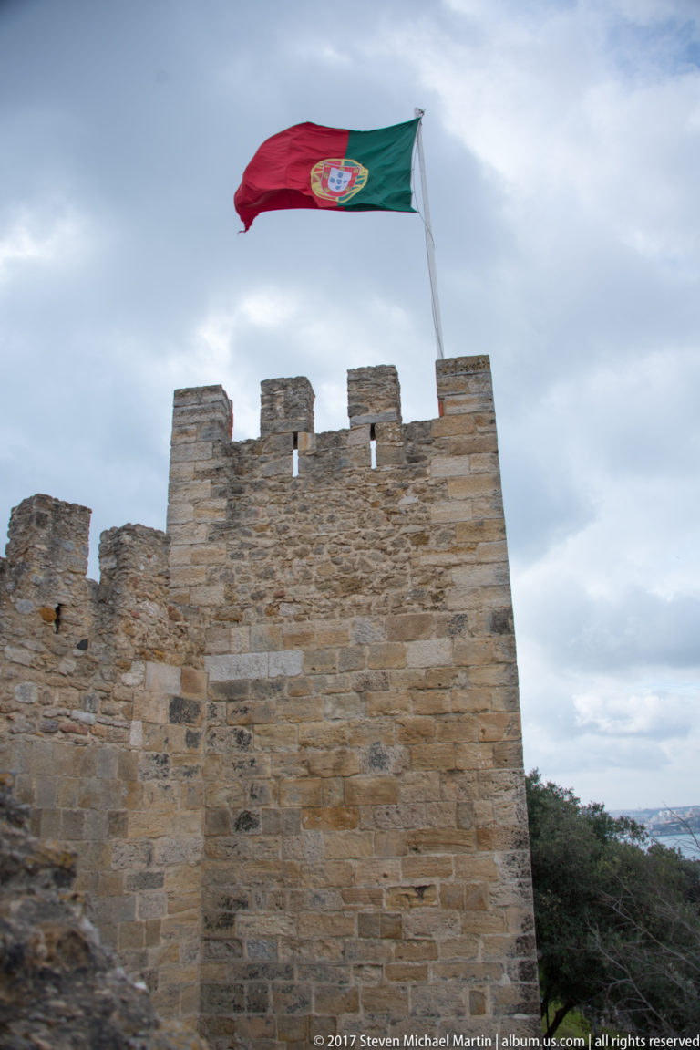 Portuguese flag flying above a square crenellated stone tower of São Jorge Castle.