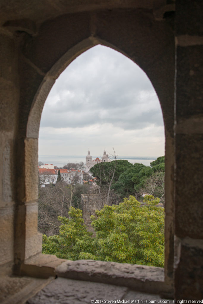 Lisbon rooftops and distant towers seen through a large rounded stone arch inside São Jorge Castle.