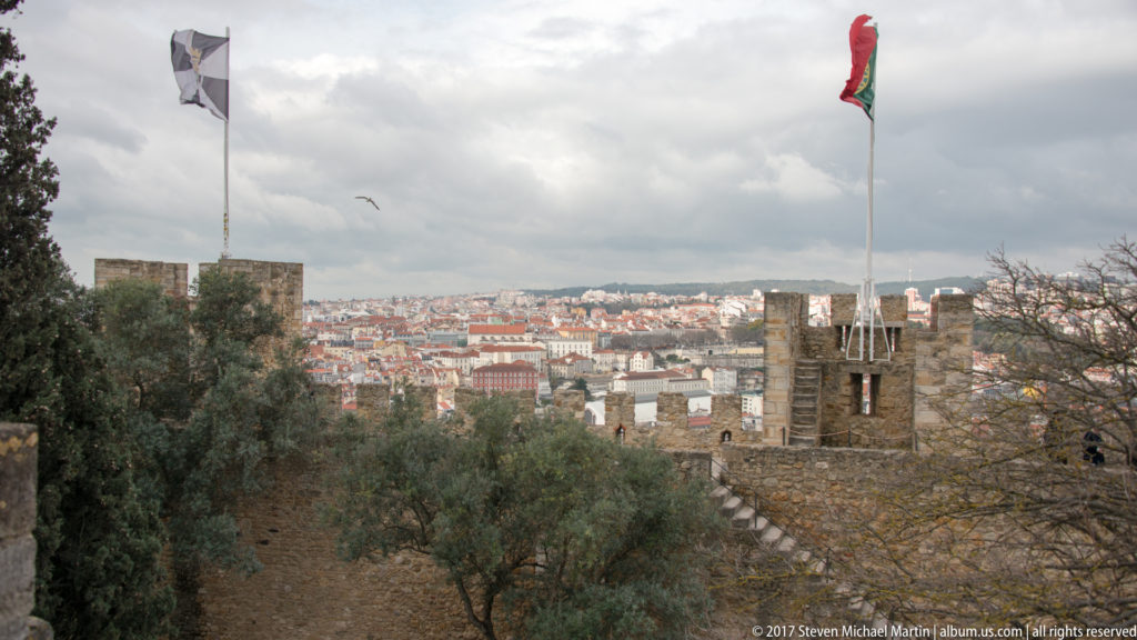 São Jorge Castle ramparts, towers, trees, and Portuguese flag overlooking the dense city of Lisbon.