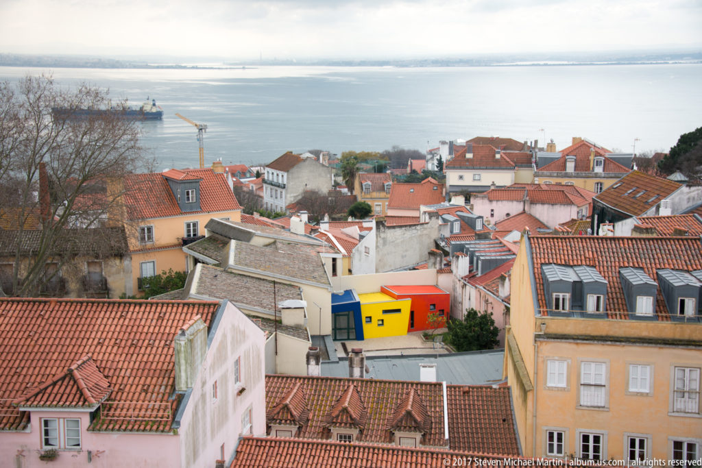Terracotta-roofed Lisbon hillside descending toward the Tagus, with bright blue, yellow, and red buildings near center.