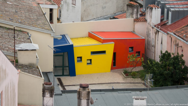 Compact modern building with vivid blue, yellow, orange, and red geometric walls amid older Lisbon rooftops.