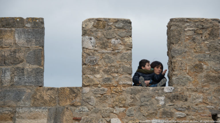 Two visitors visible through an opening between rough stone merlons on the battlements of São Jorge Castle.
