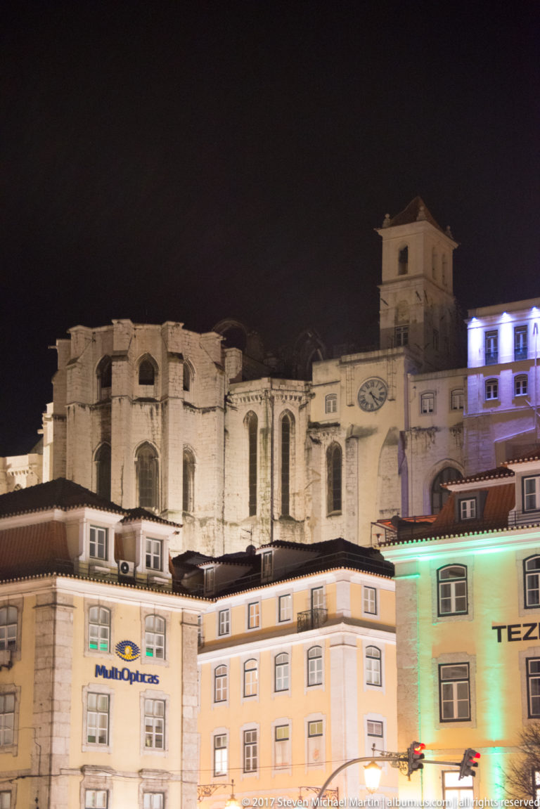Roofless stone walls and pointed Gothic openings of the Carmo Convent rising above illuminated city buildings at night.