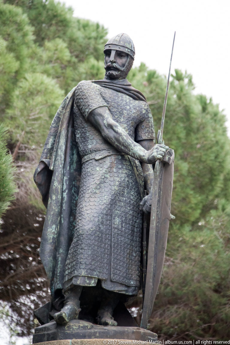 Bronze armored male figure holding a sword and shield outdoors among trees at São Jorge Castle.