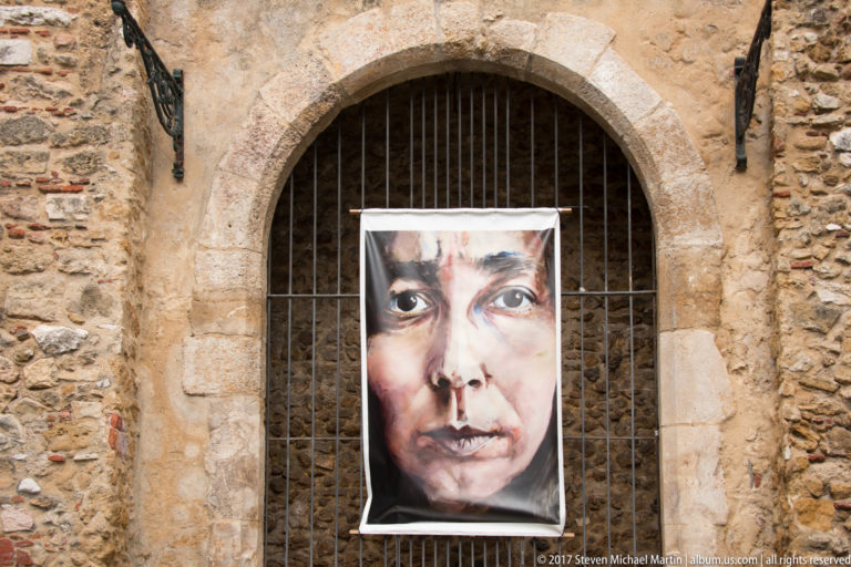 Large painted female face suspended on iron bars inside a rounded stone arch in weathered masonry.