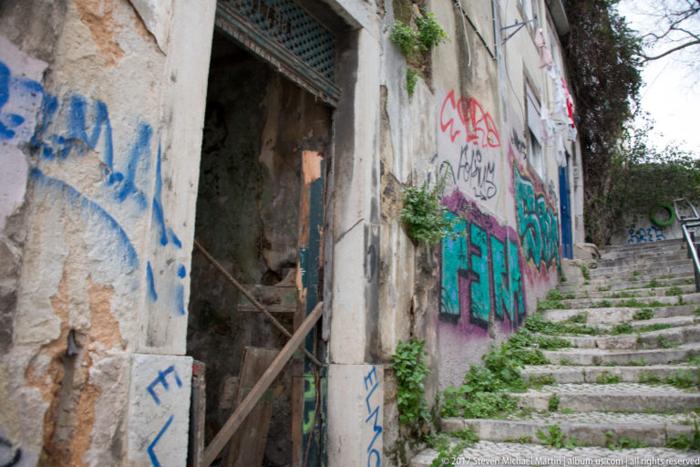 Steep stone stairway between weathered graffiti-covered buildings with railings and scattered vegetation.