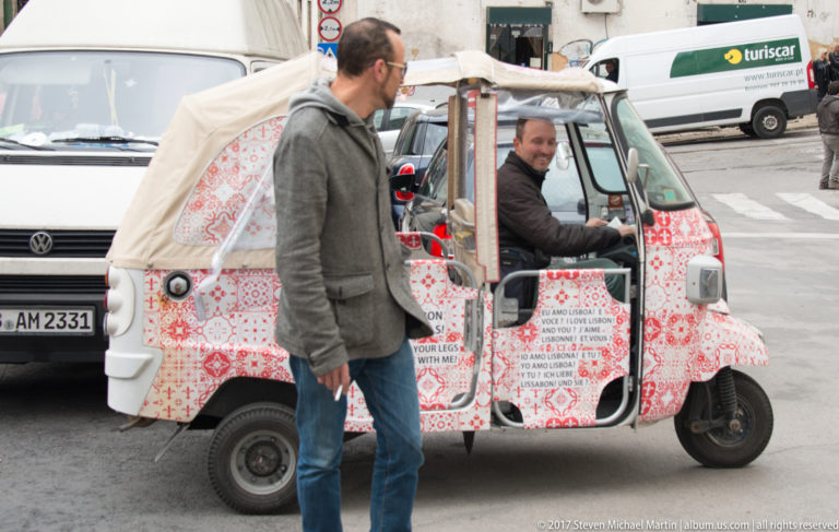 Red-and-white patterned tourist tuk-tuk with seated driver and a man standing beside it on a Lisbon street.