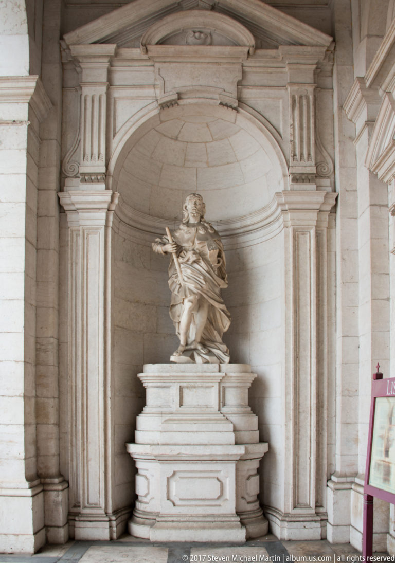 White stone statue of a robed saint gesturing outward from a deep classical architectural niche.