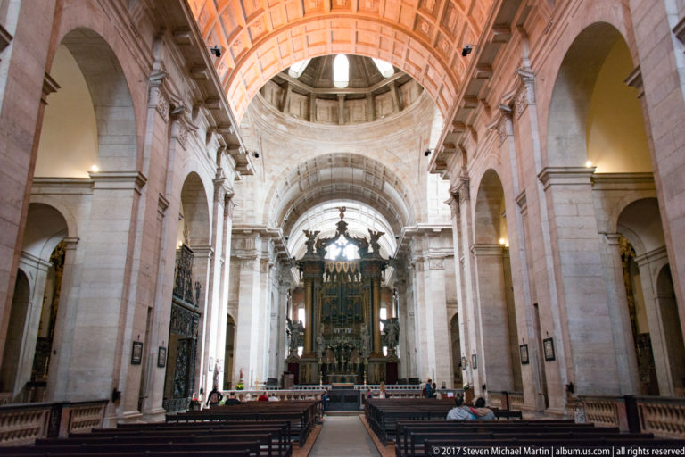 Wide nave of São Vicente de Fora with pale stone arches, coffered vault, pews, and high altar beyond.