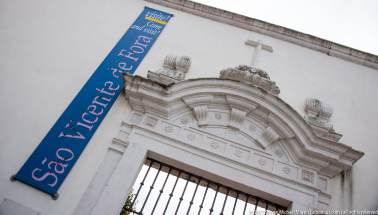 White carved entrance with cross and ornamental scrollwork beside a blue banner reading 'São Vicente de Fora.'